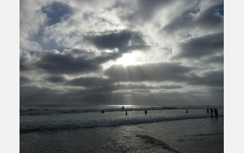 Photo of the sun peaking through clouds over a beach with people.