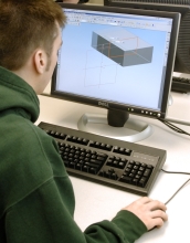 A male college student works at a computer with a 3-D image of a rectangular object on the screen.