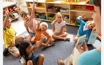 Photo of a teacher holding cards with letters and kindergarten students raising their hands.
