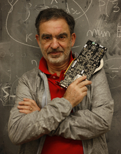 Tomaso Poggio stands in front of a blackboard holding a computer part.