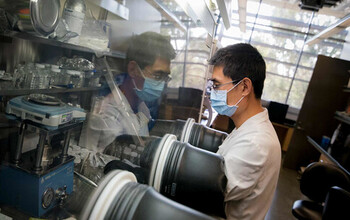 A man in a lab working on something behind a protective glass shield.