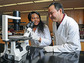 Two people in lab coats next to a microscope.