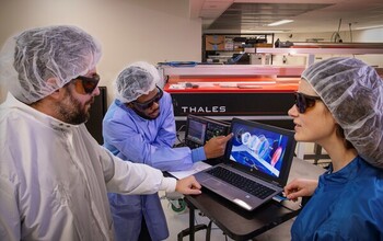 Three people wearing lab coats and goggles working in a lab with computer data.
