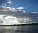 Photo of the Amazon River with rain forest on the far bank and clouds in the sky.