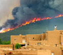Photo of a wildfire over pine forested hills in background with houses in foreground in New Mexico.