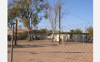 Photo through a fence of a house and sandy lot.