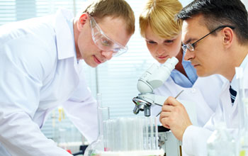 Photo of three researchers in lab coats and a microscope and test tubes on a table.