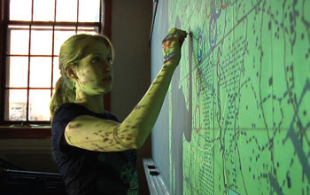 Photo of a young woman writing on a virtual green image projected onto a wall.